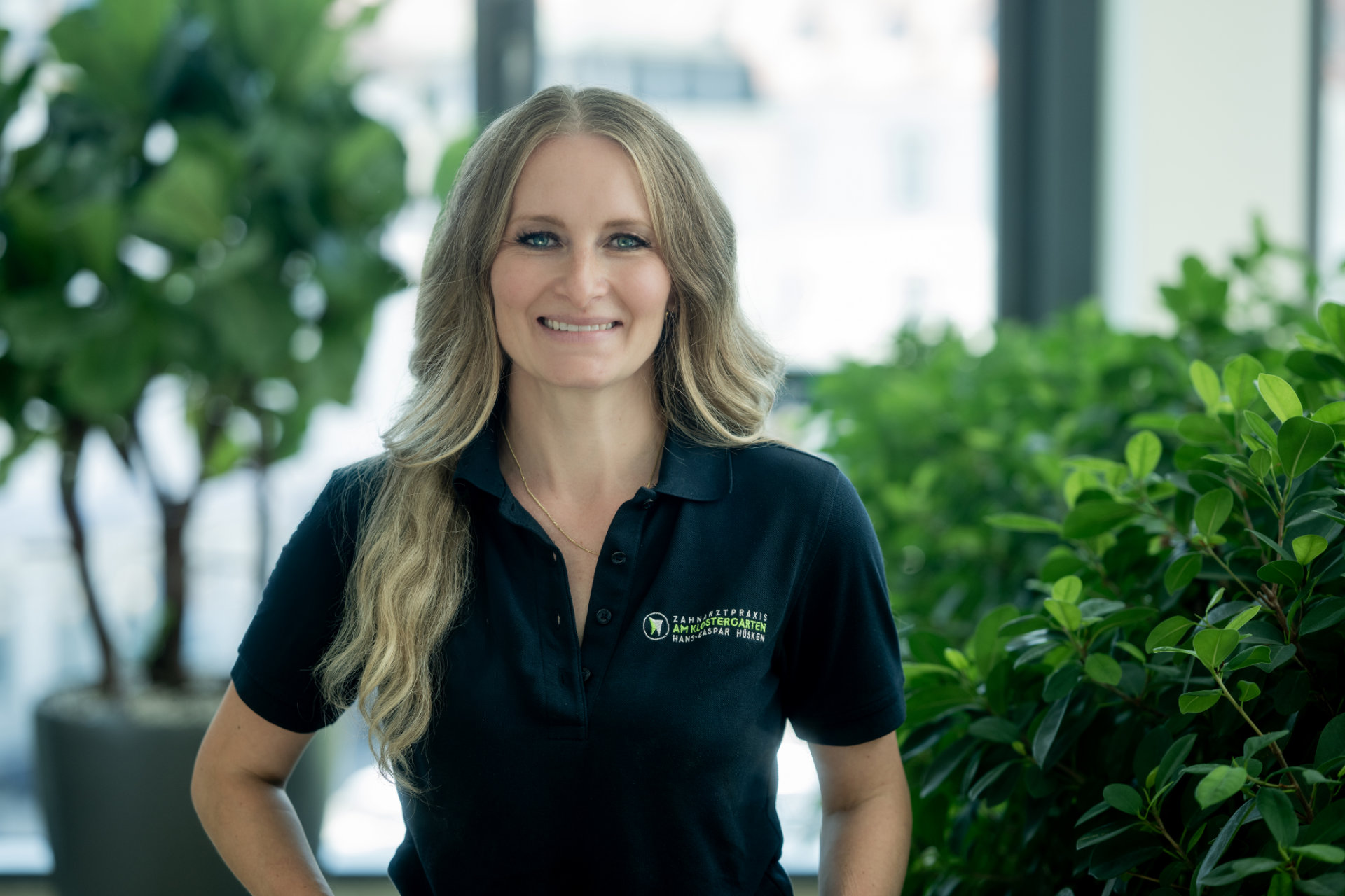 Mitarbeiterin mit langen welligen blonden Haaren vor grünen Pflanzen in der Praxis